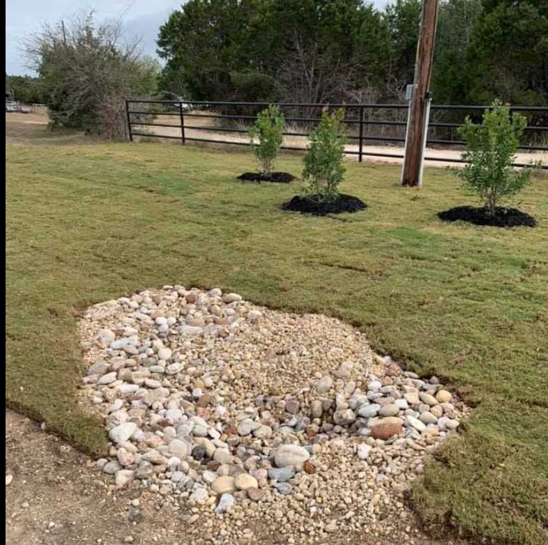 River-rock dry creek bed set into new sod with three newly planted and mulched trees behind a pipe fence