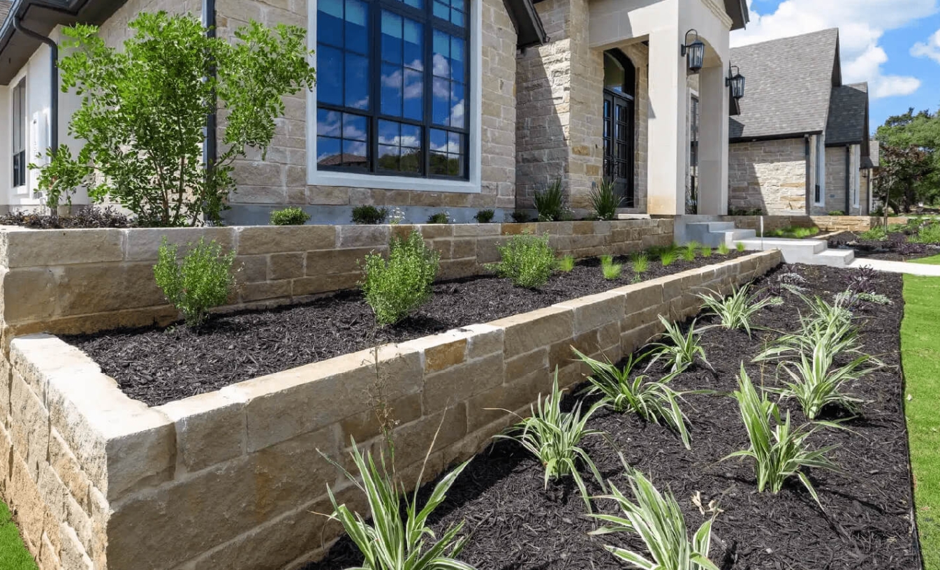 Cut limestone retaining wall holding two tiers of mulched planting beds against a stone house with black-framed windows